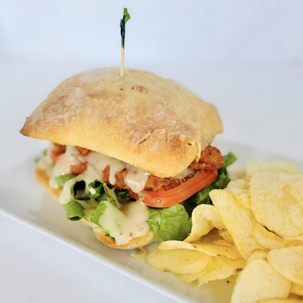 A fried chicken sandwich with lettuce, tomato, and sauce on ciabatta bread, served with potato chips.